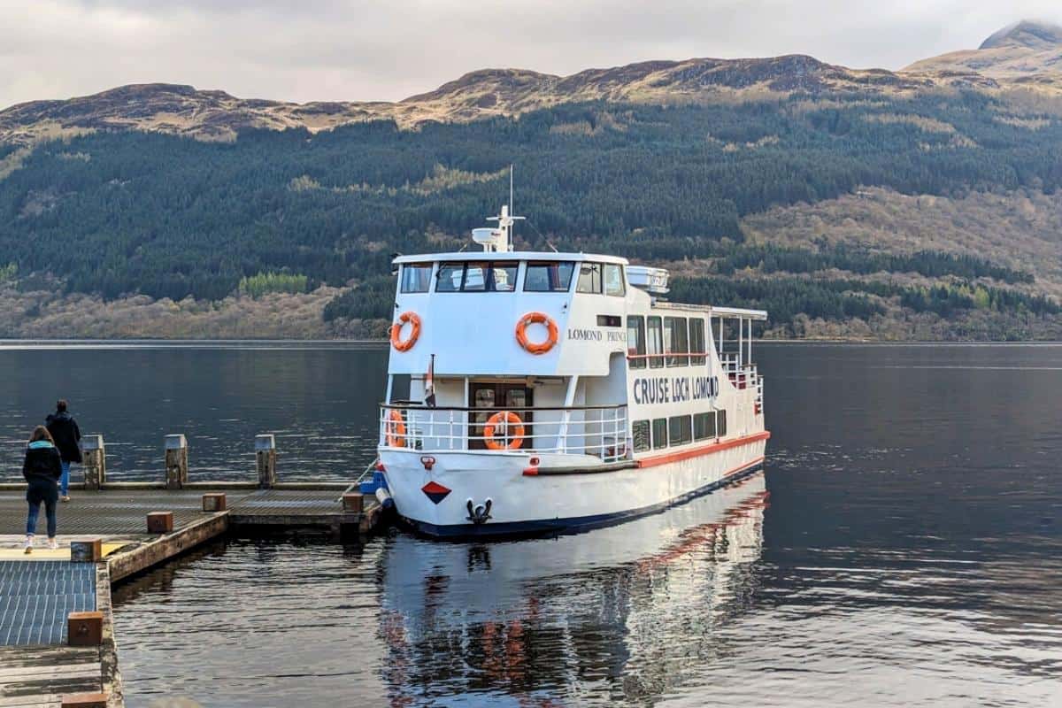 A boat is moored at the dock with the words 'Cruise Loch Lomond' on it. People are standing on the jetty and mountains are on the opposite shore. 
