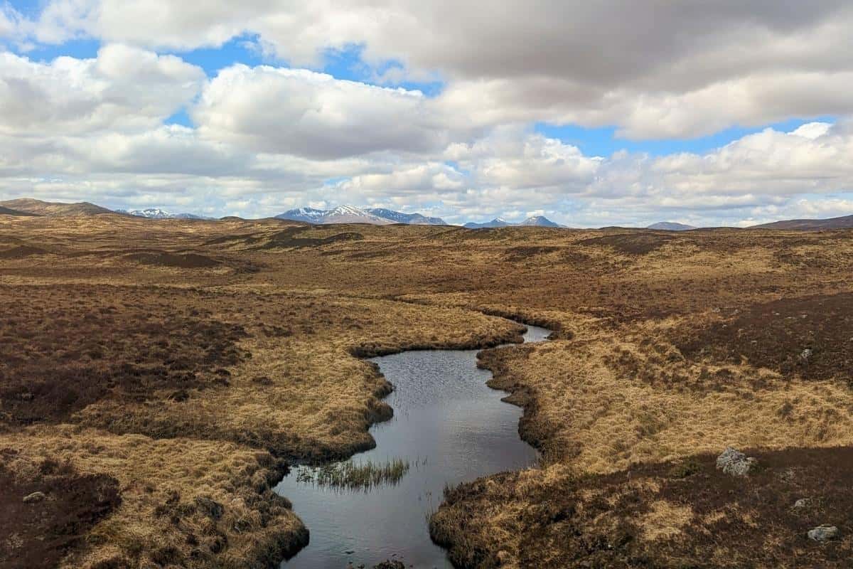7 Most Scenic Train Journeys in Scotland with Epic Views 11 Brown moorland with a river snaking through it and mountains with snow on them behind in the distance. Rannoch Moor from the West Highland Line, one of the most scenic train journeys in Scotland.