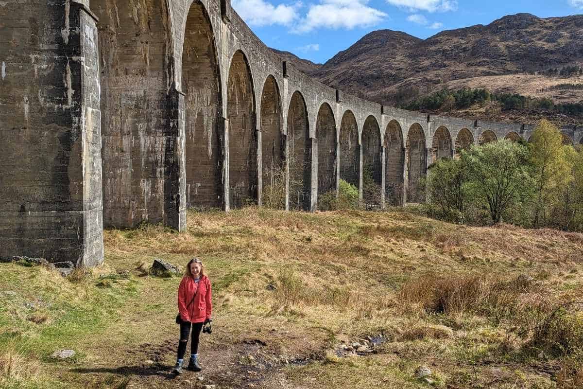Girl wearing a red jacket standing in front of a railway viaduct with rugged mountains behind. 