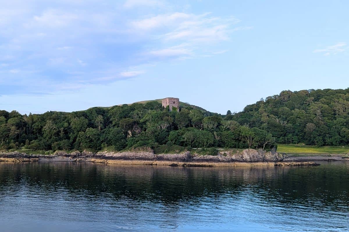 View from the water of a tree-lined coastline and a castle tower sticking up on the hill. Dunollie Castle. 