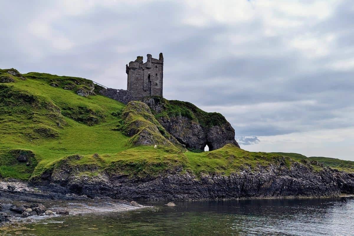 A haunting looking castle tower sits on a rocky outcrop above the water. The grass is a vibrant green. Visiting the Isle of Kerrera on a day trip is one of the best things to do in Oban. 