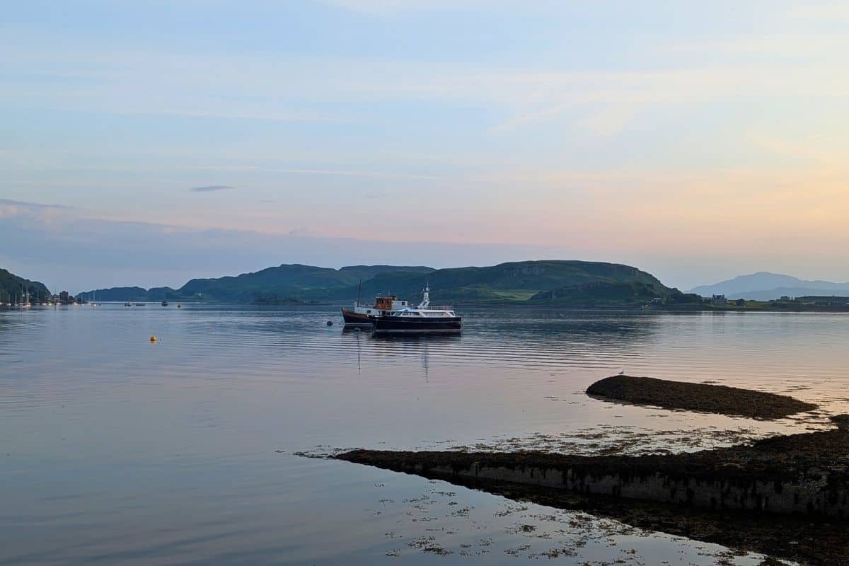Evening light falls on two moored boats with a green island behind. The water ripples peacefully. Oban at sunset. 