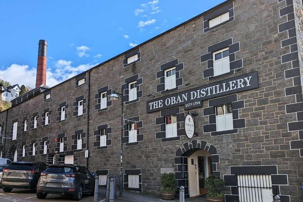 A large grey stone building with the words 'The Oban Distillery est 1974' and a large red funnel visible. 