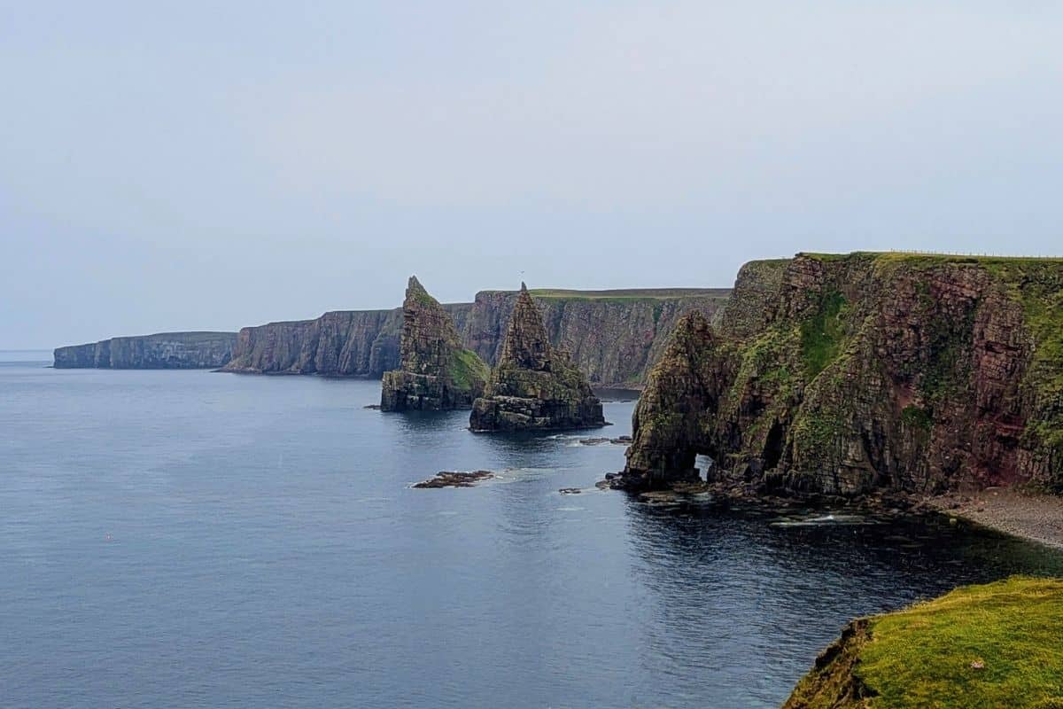 A hazy day with a vast coastline of cliffs jutting out fron the North Sea. Several needle-sharp pinnacles rise from the water. The Northern Highlands are one of the most remote Scottish regions. 