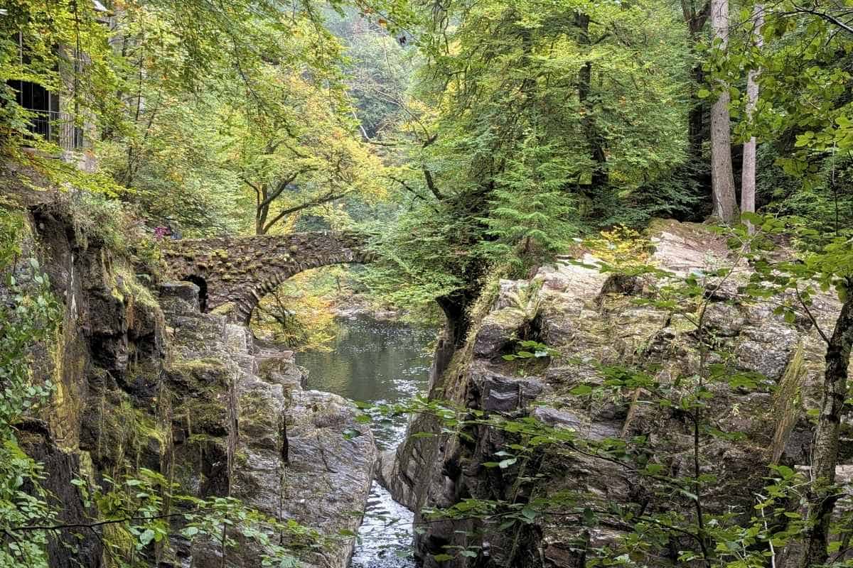 A stone bridge crosses a river surrounded by green trees starting to turn yellow. The Hermitage in Perthshire, one of the best regions in Scotland for forest walks. 