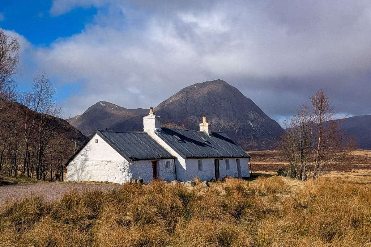 A white cottage with a black roof lies surrounded by golden grasses in front of mountains in the background. Glen Coe. 