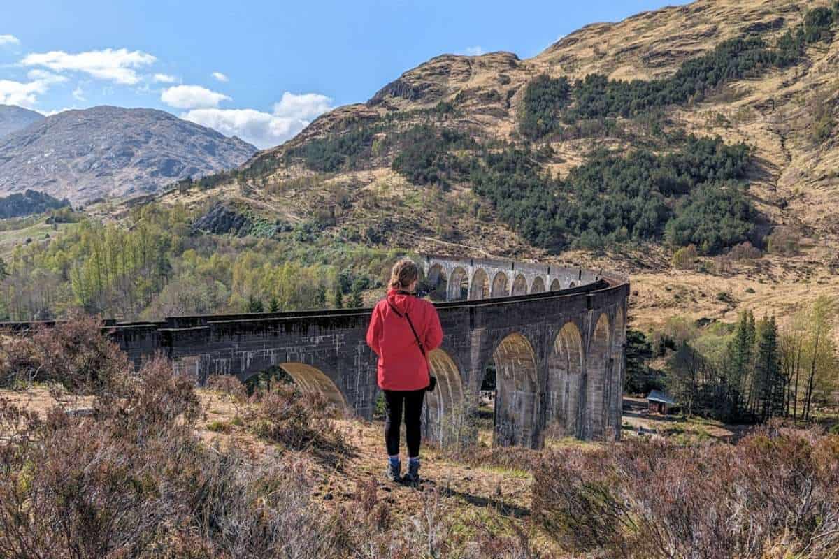 A woman wearing a red jacket stands in front of a railway viaduct surrounded by rugged mountains. Glenfinnan Viaduct. 