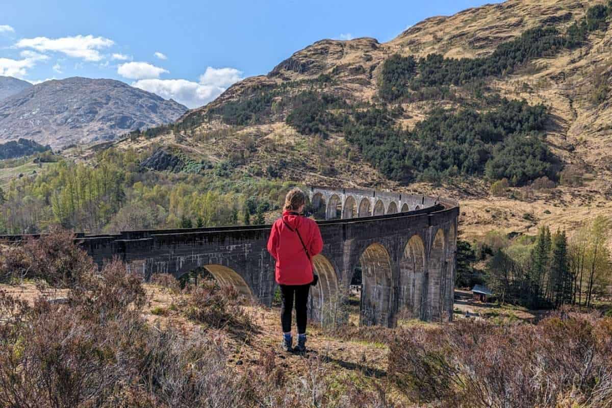 Woman wearing a red jacket standing in front of Glenfinnan Viaduct. 