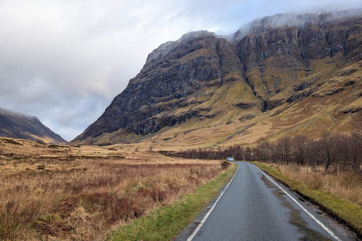 View of a rugged peak rising up with mist on top. Below it is a road with a silver car on it for this article on getting around Scotland: car vs public transport. 