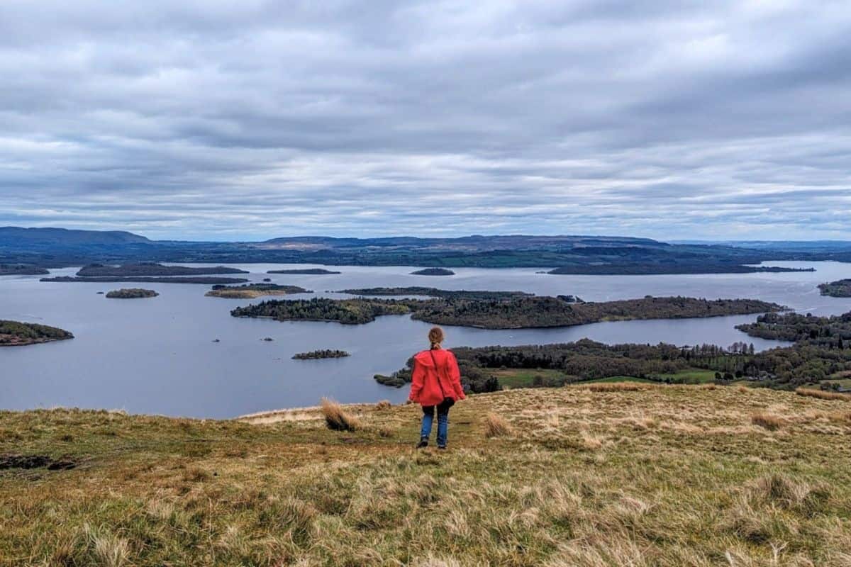A girl wearing a red jacket stands on a hill top looking down at a loch dotted with islands. Beinn Dubh for things to do in Luss. 