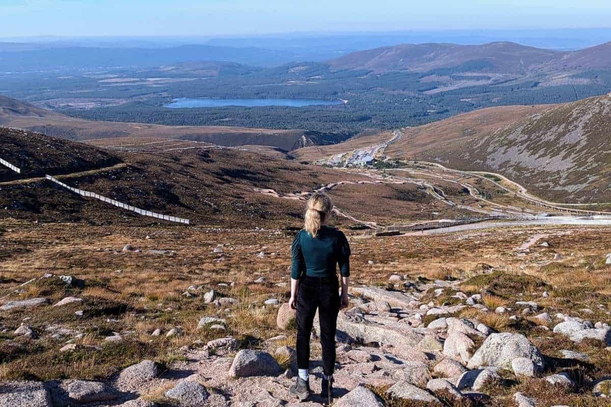 10 Easy Day Trips from Inverness Without a Car 20 A woman wearing a green top and black trousers stands looking at a viewpoint of a loch in the distance. Cairngorm Mountain is one of the best day trips from Inverness without a car.