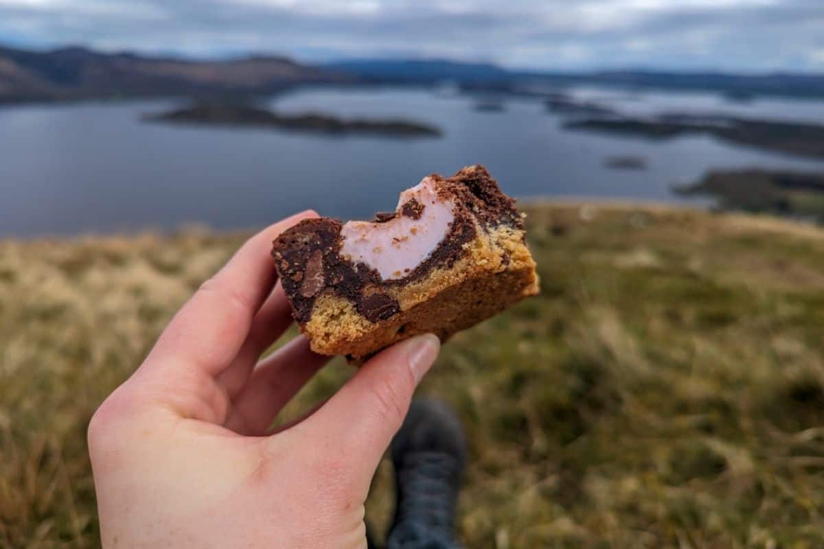 A hand holds a brookie with marshmallow in front of the blurred out scene of Loch Lomond from Beinn DUbh for things to do in Luss. 