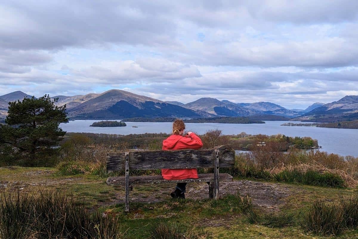 A person wearing a red jacket sits on a bench looking at the view of rugged hills and mountains surrounding a loch from Inchcailloch. 