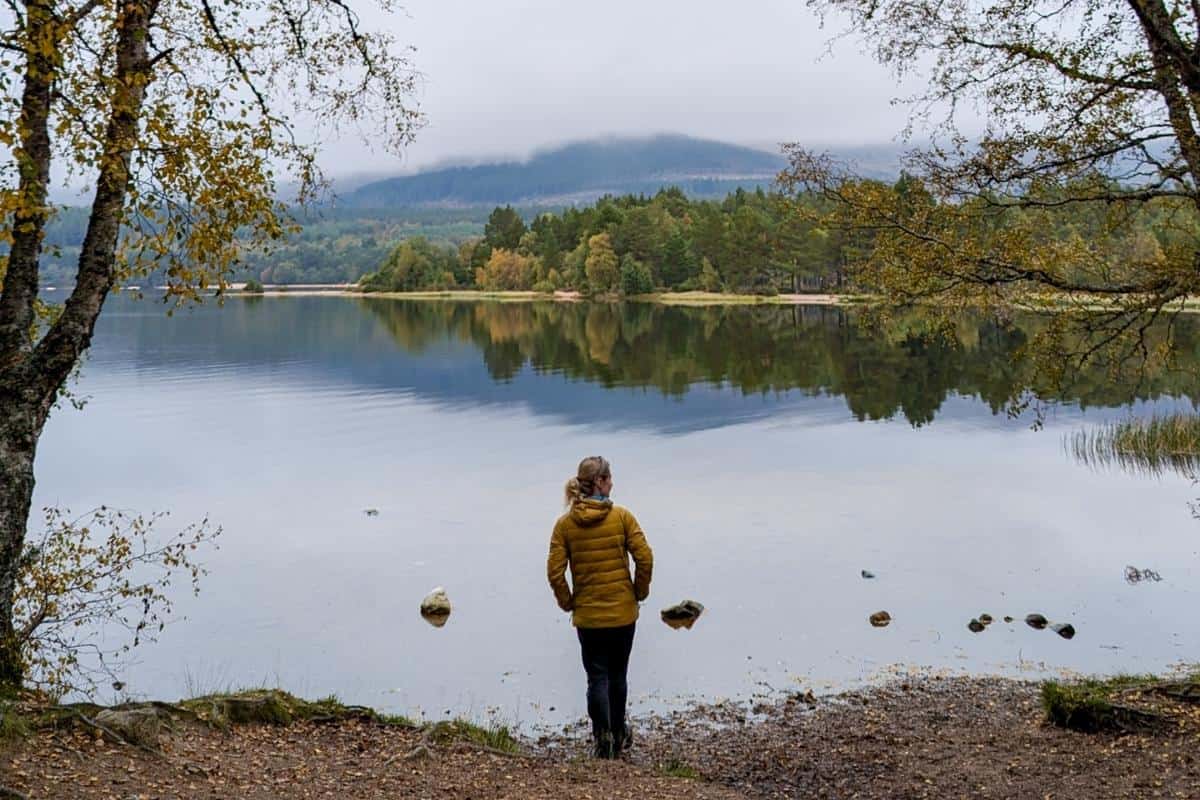 10 Easy Day Trips from Inverness Without a Car 9 A person wearing a yellow hacket stands on the loch's shore with autumn trees on the opposite bank and low-hanging cloud. Loch Morlich is one of the best day trips from Inverness without a car.
