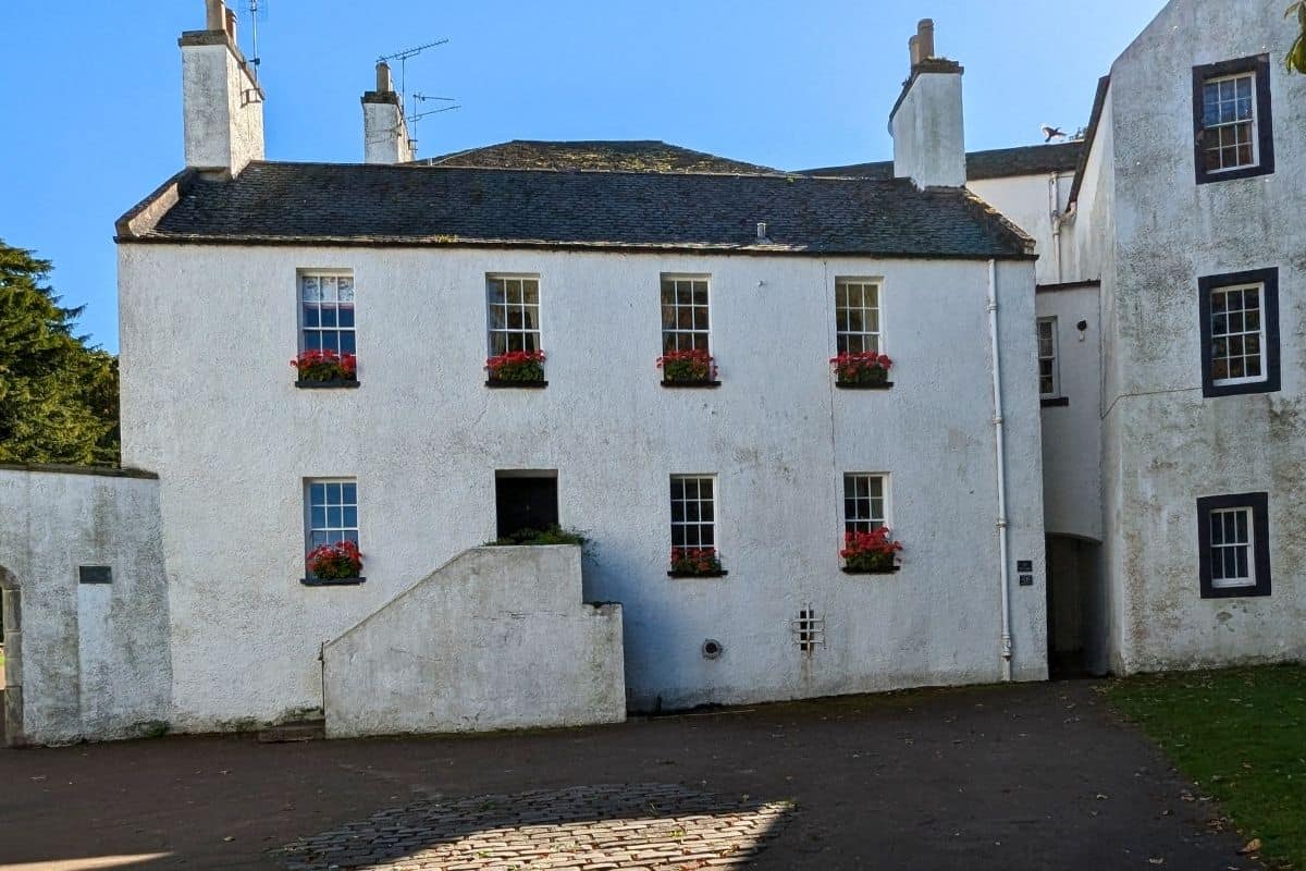 A white-washed house with a slate roof with red flowers in the window boxes. 