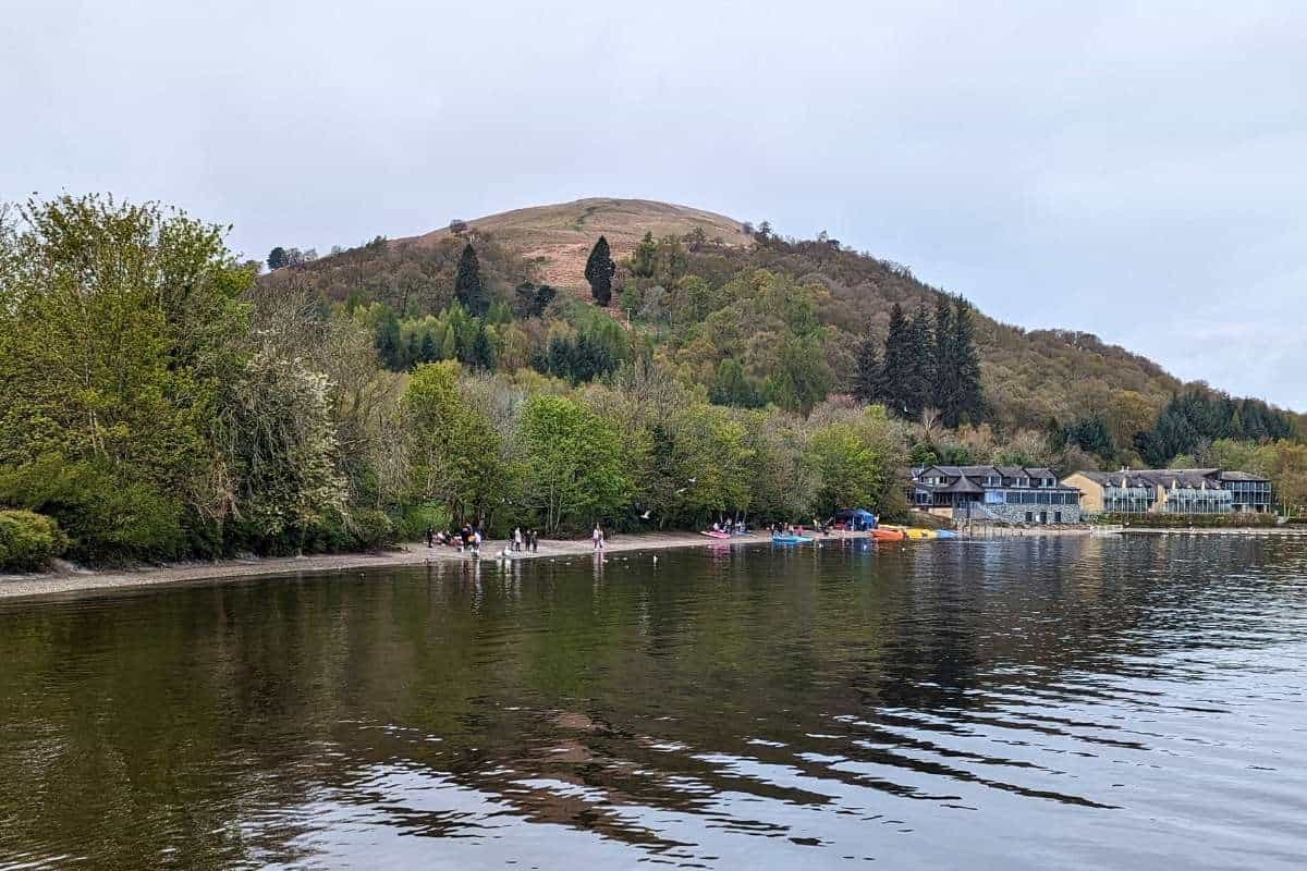 a thin beach linked with trees and a few colourful kayaks next to some buildings. Behind it a hill rises up. 