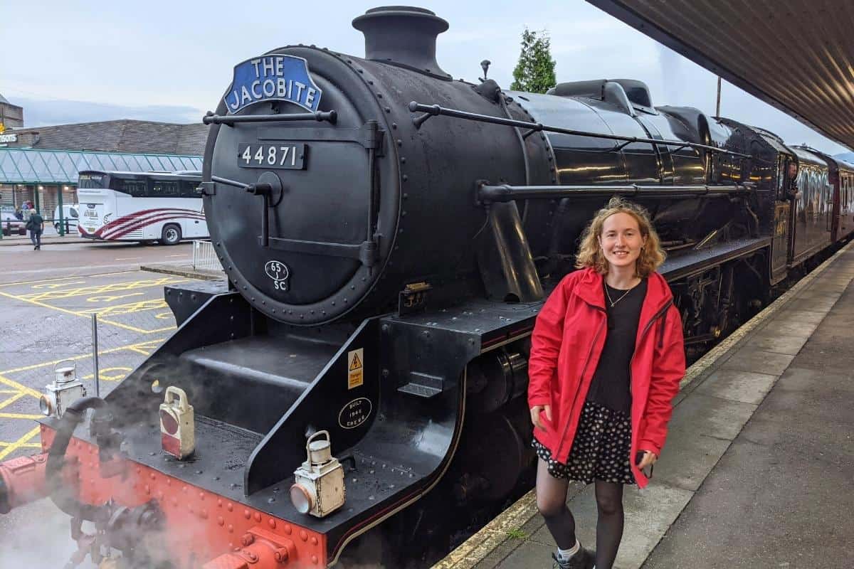 Where to Stay in Scotland Without a Car: Best Areas + Hotels 2 Little Lost Travel founder standing in front f a steam engine in Fort William. The bus station is behind.